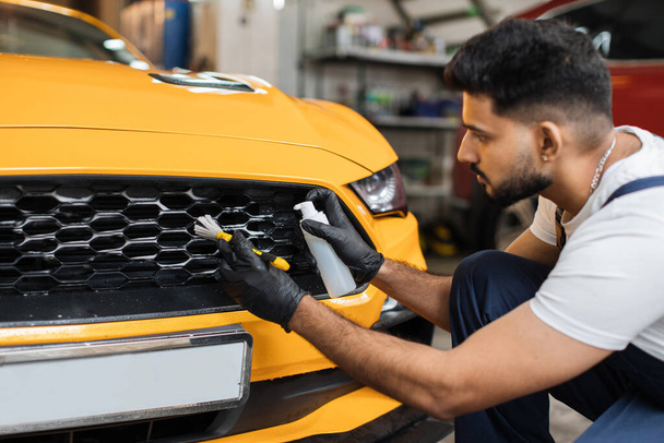 Young bearded male worker in black protective gloves and overalls cleaning radiator grille in foam with special brush. Sport yellow luxury car wash and detailing. Washing machine at the station. - Valokuva, kuva