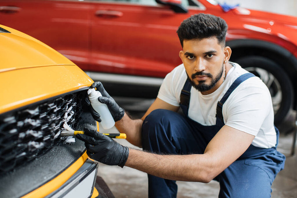 Manual cleaning with soap at car wash. Bearded male worker in rubber glove, white t-shirt and blue overalls washing car radiator grille of luxury yellow car with special brush and cleansing foam. - Foto, imagen