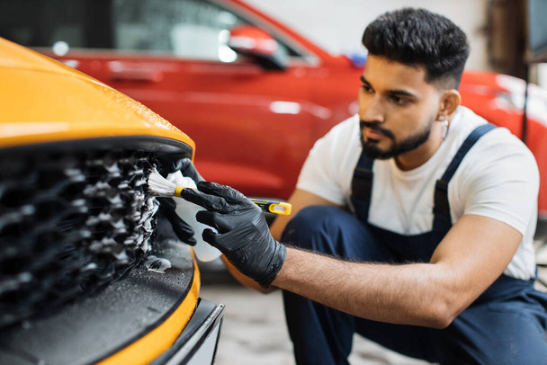 Manual cleaning with soap at car wash. Bearded male worker in rubber glove, white t-shirt and blue overalls washing car radiator grille of luxury yellow car with special brush and cleansing foam. - Foto, afbeelding