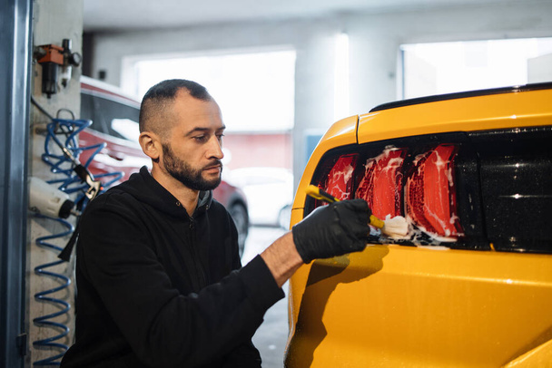 Close up photo of young male worker in black protective gloves cleaning tail light in foam with special brush. Sport yellow luxury car wash and detailing. Washing machine at the station. - Foto, Imagen