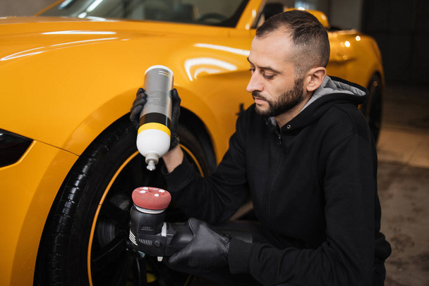 Car detailing services - polishing of yellow car. Auto service worker, wearing black clothes, puts special wax or polish cream on the orbital polisher. Auto maintenance and repair service. - Photo, image