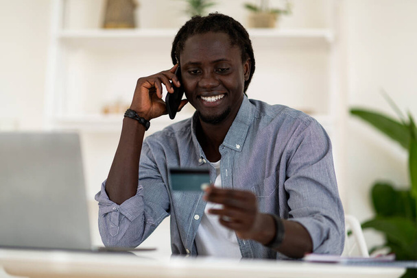 Online platby. Usmívající se černošky na volné noze Muž drží kreditní kartu a mluví na mobilní telefon, zatímco sedí na stole v kanceláři, veselý afroameričtí muži mluví s bankovním manažerem, volné místo - Fotografie, Obrázek