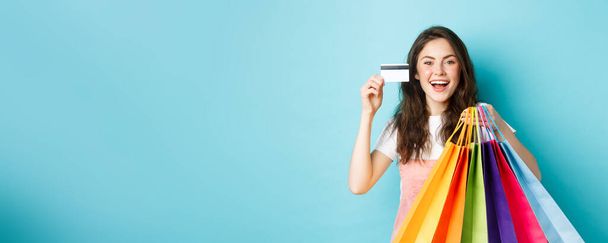 Glückliche schöne Frau zeigt Plastikkreditkarte und Einkaufstüten mit Waren, kauft mit Rabatten, steht vor blauem Hintergrund. - Foto, Bild