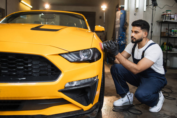 Car detailing and polishing concept. Professional young male car service worker in uniform, holding in hands orbital polisher, and polishing yellow luxury car at auto repair service station. - Photo, image