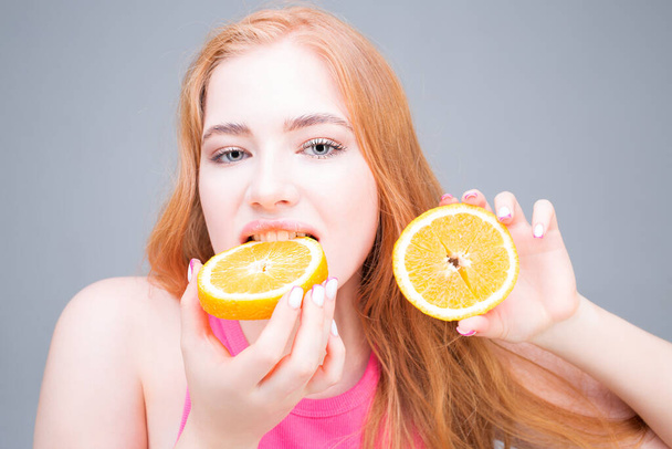 幸せな若い美しい女性が灰色の背景に隔離されたオレンジのジューシーな半分を食べる。健康的な食事の概念。国会. - 写真・画像