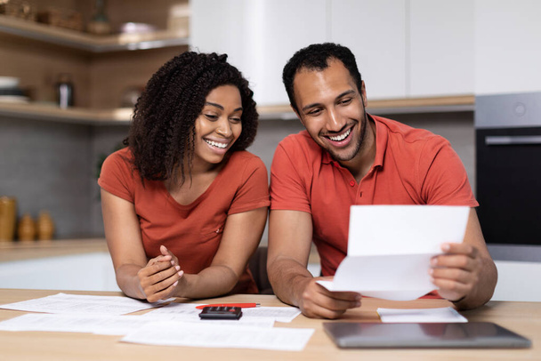 Happy young black lady in red t-shirt shows documents to guy with computer at kitchen interior. Great news, bill payment, home bookkeeping, taxes together, household chores, end of mortgage and credit - Zdjęcie, obraz