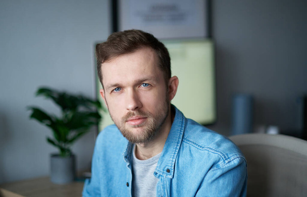 Young handsome man in denim shirt, positive emotion with computer monitor on background. Cheerful bearded male sitting in home office looking at camera with serious emotion. High quality image - Fotografie, Obrázek