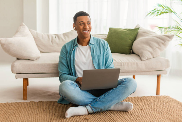 Cheerful millennial african american guy in wireless headphones with laptop looking at empty space in cozy living room interior. Video call, study and work at home, freelancing remotely, ad and offer - Photo, Image