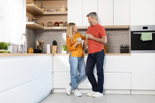 Szczęśliwa romantyczna para w średnim wieku atrakcyjny mężczyzna i kobieta stojący przy stole kuchennym w domu, trzymający kubki, pijący kawę, rozmawiający i uśmiechający się, panorama z przestrzenią do kopiowania, ujęcie full length - Zdjęcie, obraz