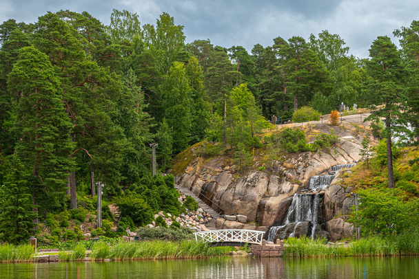 Finnország, Kotka - 2022. július 18.: Sopokanlahti park és tó. Oldalnézet a vízesés sziklák felett meg zöld lombozat környezetben kis fehér fa gyalogos híd. Szürke felhő - Fotó, kép
