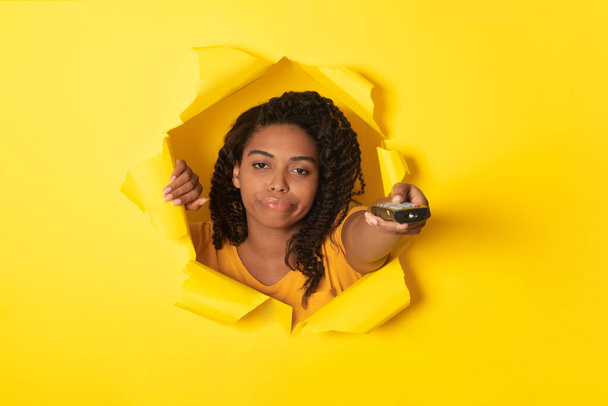 Tyytymätön Afrikkalainen Amerikkalainen nainen katselee TV-kanavat osoittava kaukosäädin reiän läpi repeytynyt keltainen paperi, Studio Shot. Tylsä elokuvakonsepti. Kollaasi - Valokuva, kuva
