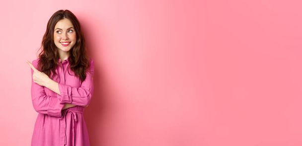 Menina glamour bonita com maquiagem, vestindo roupa de primavera, apontando e olhando para o logotipo, mostrando banner no espaço livre esquerdo, de pé contra fundo rosa. - Foto, Imagem