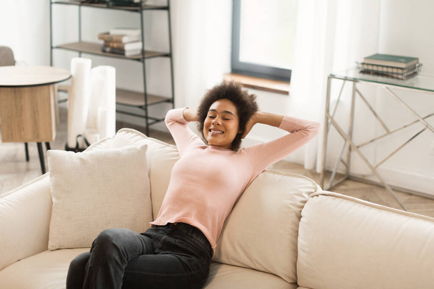 Szczęśliwy zrelaksowany dość młoda Afroamerykanka z zamkniętymi oczami siedzi na kanapie, cieszyć się ciszą, komfort w jasnym nowoczesnym salonie wnętrza, wolnej przestrzeni. Przerwa i odpoczynek we własnym domu, covid-19 - Zdjęcie, obraz