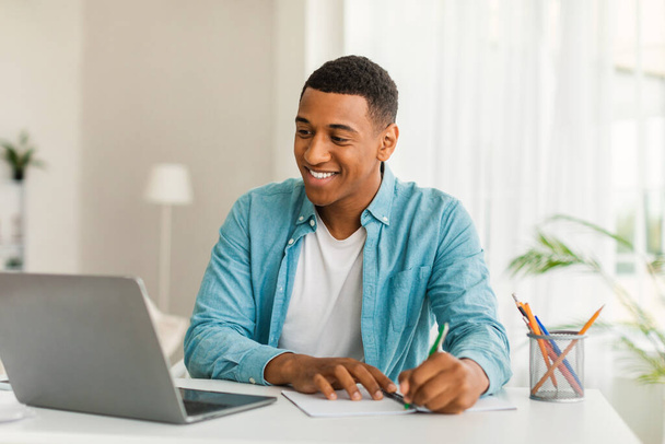 Glad handsome young african american man manager in casual use computer for work, study, making notes in room or office interior. Business and freelance remotely, social distance at home due covid-19 - Photo, image