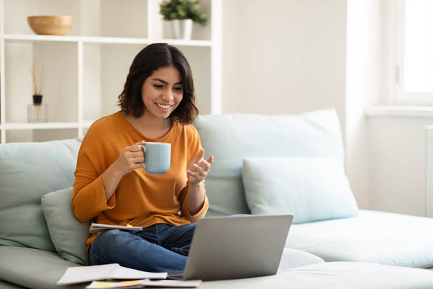 Διαδικτυακή επικοινωνία. Ευτυχισμένη αραβική γυναίκα κάνει βιντεοκλήση στο Laptop και πίνοντας καφέ στο σπίτι, χαρούμενη γυναίκα της Μέσης Ανατολής έχοντας τηλεδιάσκεψη με τους φίλους και απολαμβάνοντας ζεστό ποτό, αντίγραφο χώρου - Φωτογραφία, εικόνα