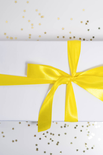 vertical photo of holiday gift with yellow satin ribbon closeup on white background with sparkling confetti stars - Photo, Image
