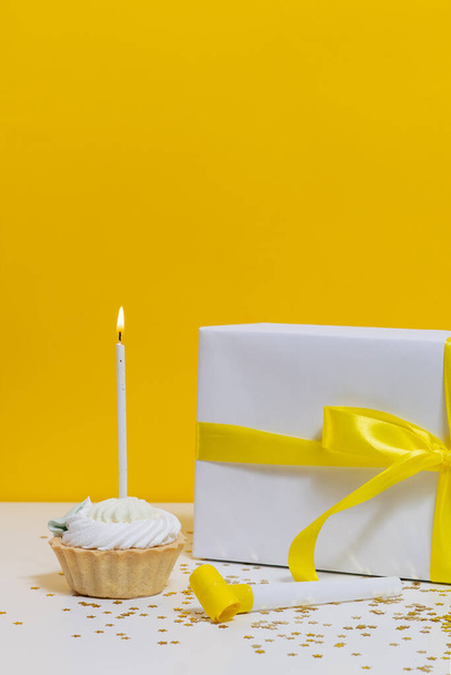 Függőleges felvétel egy ünnepi muffinról, egy gyertyával, ajándékkal és egy partikürttel az asztalon, arany konfettivel díszítve, csillagok formájában, sárga, elszigetelt háttérrel. ötlet az ajándék, születésnapi ünneplés - Fotó, kép