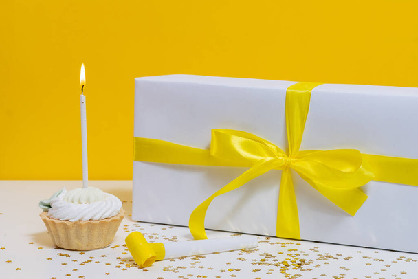 Narozeninový dortík s jednou hořící svíčkou, houkačkou a dárkem leží na stole s třpytivými konfety na žlutém izolovaném pozadí. horizontální pohled. dovolená koncept a dárek nápad - Fotografie, Obrázek