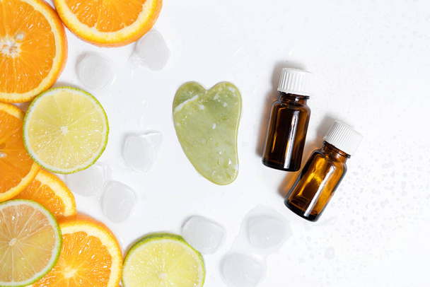 bocaux en verre brun d'huile et un massage guasha se trouvent sur fond blanc avec de la glace et des agrumes sous la forme de citron vert et orange - Photo, image