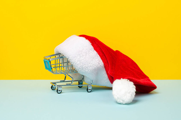Grocery cart from the supermarket in a Santa Claus hat isolated on yellow background. New year sale concept with red Santa hat and shopping trolley - Fotoğraf, Görsel