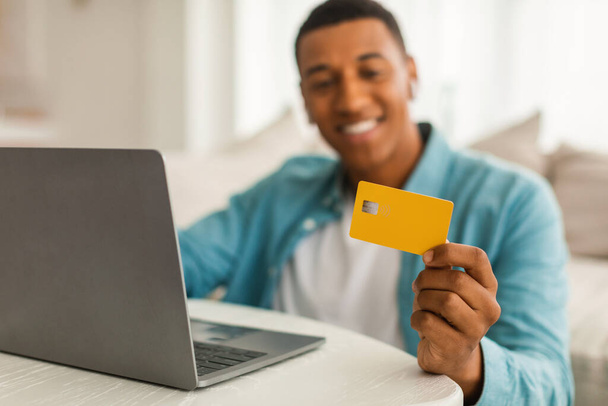 Ciesz się tysiącletniego afrykańskiego mężczyzny patrząc na kartę kredytową z laptopem, cieszyć się zakupy online w salonie wnętrza, zamazane, zbliżenie. Zakupy w domu, ogromna sprzedaż, finanse i bankowość zdalnie - Zdjęcie, obraz