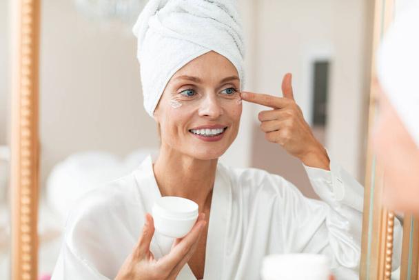 Mooie middelbare leeftijd vrouw in zijdezachte badjas aanbrengen hydraterende oogcrème, kijken naar spiegel, genieten van gezichtsverzorging routine, badkamer interieur - Foto, afbeelding