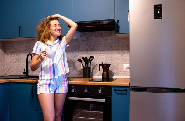 Portrait d'une jolie jeune femme en pyjama tenant une tasse et fixant ses cheveux dans la cuisine. Souriant, regardant par la fenêtre. Photo de haute qualité - Photo, image