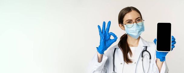 Online medical help concept. Enthusiastic young woman doctor in face mask, showing okay sign and mobile phone app, smartphone screen interface, white background. - Foto, afbeelding