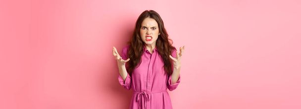 Angry and furious young woman shaking hands, looks frustrated and outraged, arguing, complaining on something, standing against pink background. - Photo, Image