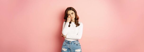 Image of sad and bored, reluctant young woman looking at smth boring, feeling lonely, leaning head on fist with indifferent face expression, standing over pink background. - Фото, изображение