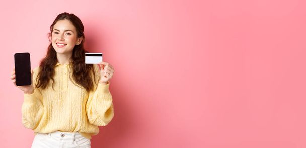 Tecnología y compras en línea. Retrato de la hermosa dama que muestra la pantalla del teléfono móvil y la tarjeta de crédito de plástico, sonriendo complacido, recomiendo aplicación, de pie sobre fondo rosa. - Foto, Imagen