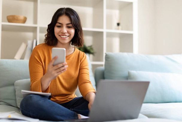 Νεαρή γυναίκα της Μέσης Ανατολής χαλαρώνοντας στον καναπέ με Smartphone και Laptop στο σπίτι, χαμογελώντας Millennial Arab Woman Browsing Νέα εφαρμογή στο κινητό τηλέφωνο και περιήγηση στο Internet στον υπολογιστή, Αντιγραφή χώρου - Φωτογραφία, εικόνα