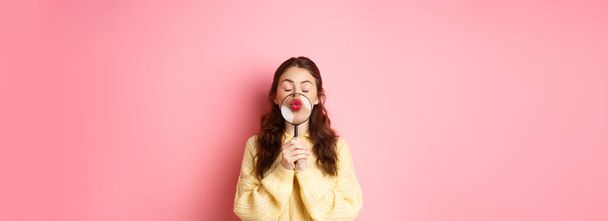 Jeune femme mignonne et romantique magnifier ses lèvres avec une loupe, faire visage baisers, debout avec les yeux fermés sur fond rose. - Photo, image