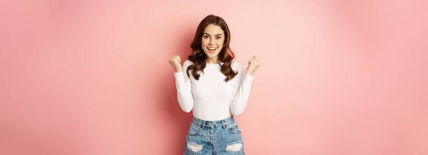 Enthusiastic brunette girl clench fists and smiling excited, feeling empowered and confident, standing cheerful against pink background. - Fotografie, Obrázek