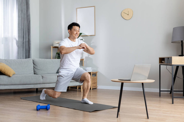 Motivated athletic handsome middle aged asian man exercising at home, training on yoga mat, using laptop, watching online fitness class, copy space. Workout plan for men concept - Photo, Image