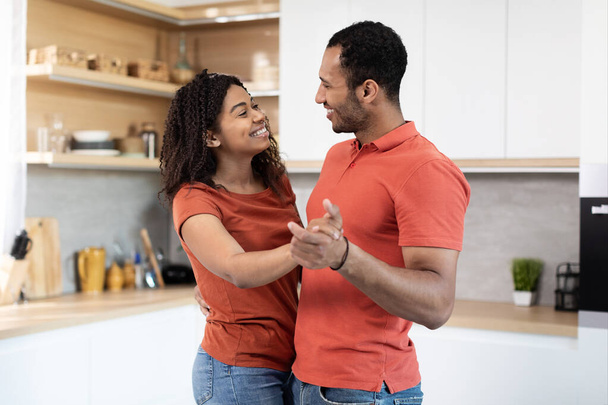Smiling handsome young black husband and wife in red t-shirts dancing, enjoy free time and music, have fun together at kitchen interior. Anniversary, date, love, romance and relationships at home - Photo, Image
