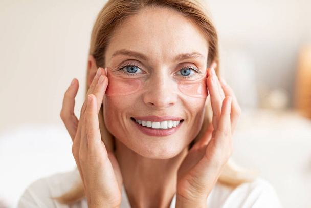 Happy mature lady wearing cosmetic eye patches and smiling at camera, making rejuvenating procedure, pampering herself, doing daily skincare routine at home - Photo, Image