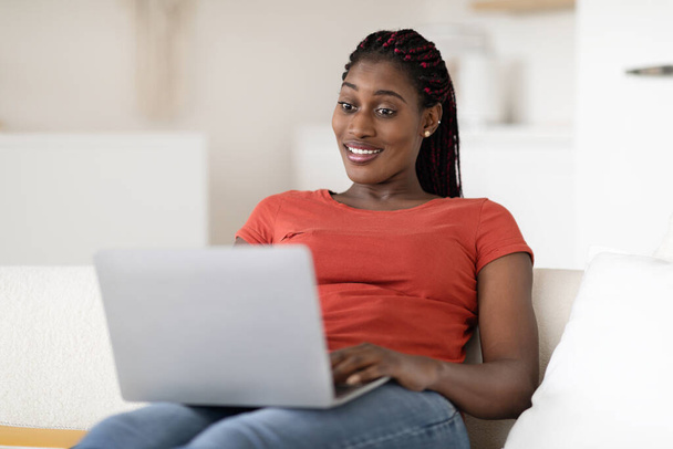 Online communicatie. glimlachende jonge zwarte vrouw zitten op de bank en met behulp van laptop, gelukkig Afro-Amerikaanse vrouw messaging met vrienden in het internet of werken op afstand als freelancer, kopiëren ruimte - Foto, afbeelding