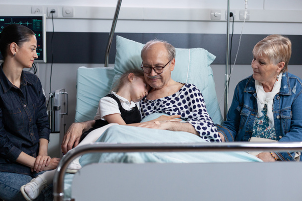 Little girl hugging grandfather at sanatorium room before surgery in medical tower. Hospitalized old man saying goodbye to close family members at elderly people hospital. - Photo, Image