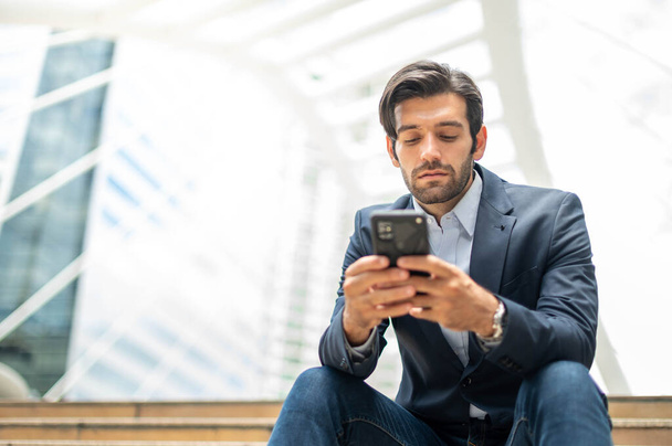 Close-up de um jovem caucasiano homem sério usando um dispositivo de smartphone moderno enquanto sentado em uma área pública, enquanto olha para a rede social em seu smartphone. - Foto, Imagem