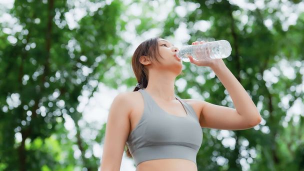 Young asian woman drinking water from a bottle while taking a break during her workout. Happy adolescence female working out in the park and taking rest a break to drink water - Photo, image