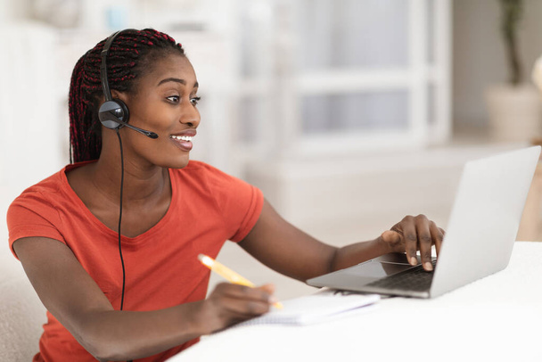 Fernstudium. Lächelnde Schwarze im Headset Online-Studium mit Laptop zu Hause, glückliche Afroamerikanerin, die Webinar schaut und Notizen zum Notizblock macht, Remote-Bildung genießt, freien Raum - Foto, Bild