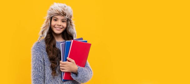 happy kid long hair in hat with copybook on yellow background, education. Banner of child girl in winter hat, studio poster header with copy space - Photo, Image
