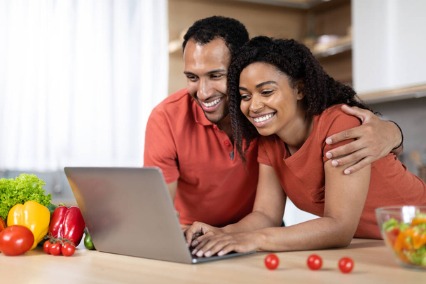 彼の妻を抱きかかえた赤いTシャツの幸せな若い黒人男性は、キッチンのインテリアで有機野菜とテーブルでコンピュータ上でビデオ通話をしています。フードブログ,遠隔会議, covid-19流行による通信 - 写真・画像