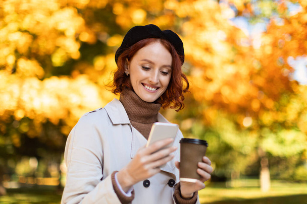Uśmiechnięta młoda, ruda kobieta w płaszczu przeciwdeszczowym, czytająca wiadomości na smartfonie, pijąca drinki na wynos w parku miejskim jesienią. Nowa aplikacja, blog i czat w sieciach społecznościowych outdoor, przerwa na kawę - Zdjęcie, obraz