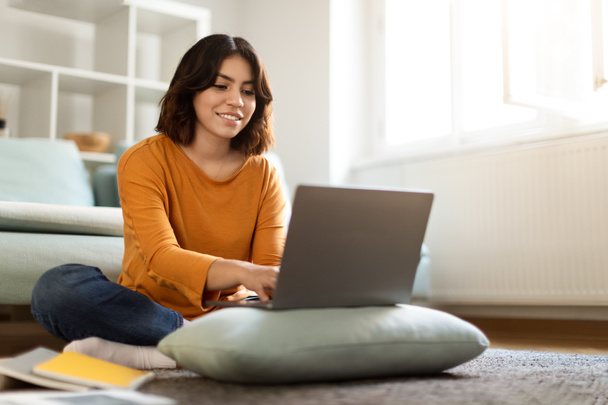 Online koulutus. Hymyilevä nuori arabinainen tutkimus kannettavan tietokoneen kanssa kotona, onnellinen Lähi-idän naispuolinen opiskelija kirjoittaa tietokoneen näppäimistöllä, kun hän istuu lattialla huoneessa, nauttien etäopiskelusta - Valokuva, kuva