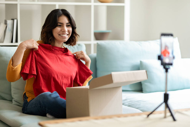 Beautiful Young Arab Woman Making Unboxing Video For Her Fashion Vlog, Smiling Middle Eastern Woman Unpacking Parcel With New Clothes In Front Of Camera At Home, Using Smartphone On Tripod - 写真・画像