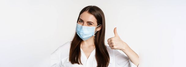 Close up portrait, face of woman in medical mask smiling, showing thumbs up, standing over white background. Copy space - 写真・画像