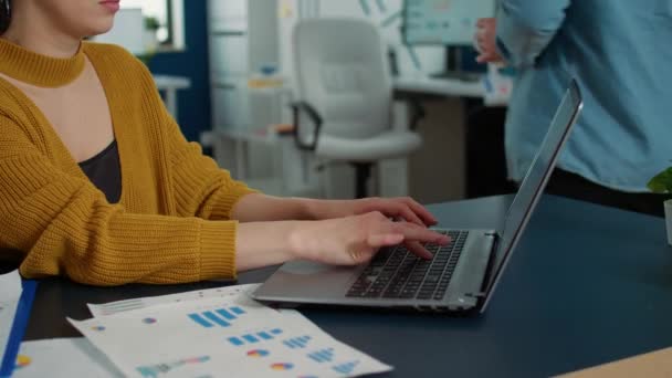 Portrait of woman typing using laptop keyboard and smiling at camera sitting at desk in busy startup office. Casual business employee working relaxed with papers with charts and statstics. - Filmagem, Vídeo