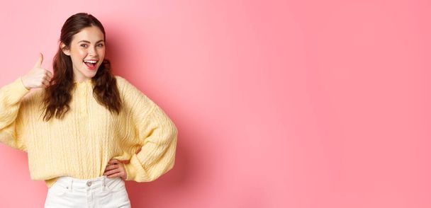 Ritratto di bella bruna modello femminile mostrando pollici in su, come la tua idea, dare supporto, approvare o lodare qualcosa di buono, in piedi su sfondo rosa. - Foto, immagini